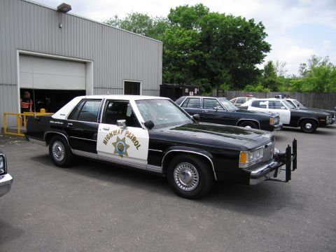 1991 Ford LTD Crown Victoria - black-and-white Ford Highway Patrol sedan with a gold star shield is parked outside a metal warehouse among other cruisers.