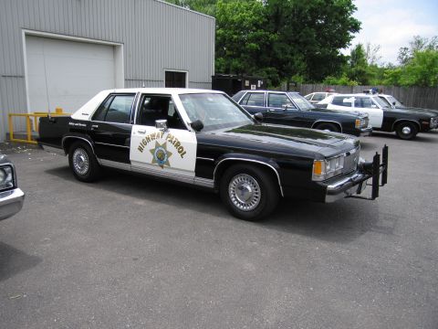 1991 Ford LTD Crown Victoria - black-and-white Ford Highway Patrol sedan with a gold star shield and push bar is parked in a lot with other vintage cruisers behind.