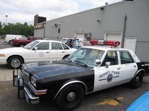 1991 Ford LTD Crown Victoria - black-and-white sedan marked JAFFREY POLICE with a red rotating roof beacon is parked outside a metal warehouse beside other cars.