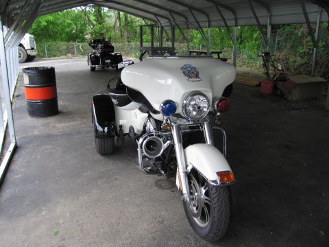 1991 Ford LTD Crown Victoria - white police-style Harley-Davidson trike with a windshield and emblem sits under a carport beside a black motorcycle.