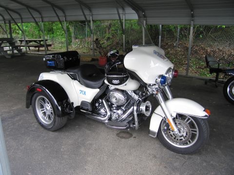 1991 Ford LTD Crown Victoria - white touring trike marked 701 with a helmet resting on its seat sits under a metal carport near a picnic table.