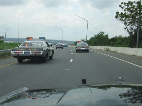 1991 Ford LTD Crown Victoria - black-and-white Highway Patrol sedan with roof lightbar leads other police cars down a multi-lane highway.
