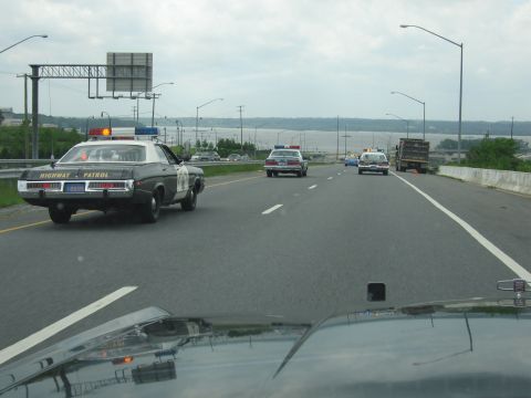 1991 Ford LTD Crown Victoria - black-and-white Highway Patrol cruiser leads a line of vintage police cars along a highway beside a body of water.