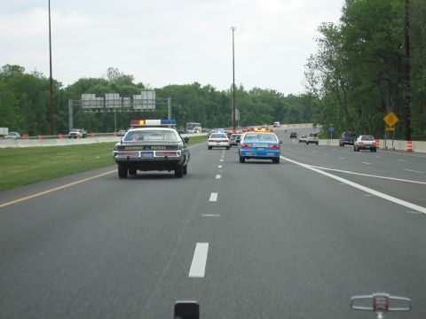 1991 Ford LTD Crown Victoria - Highway Patrol cruiser and a blue Police car drive abreast leading other vehicles on a divided highway.