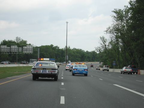 1991 Ford LTD Crown Victoria - black-and-white Highway Patrol car and a blue Police car lead traffic on a highway lined with trees.