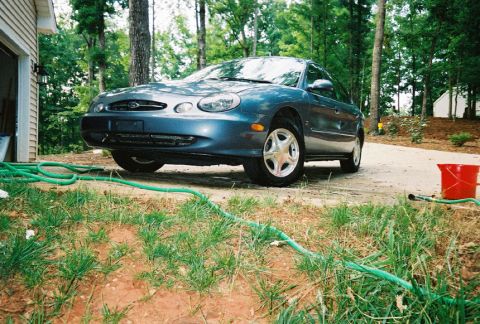 1999 Ford Taurus - A dark blue sedan is parked in a residential driveway surrounded by trees and grass.
