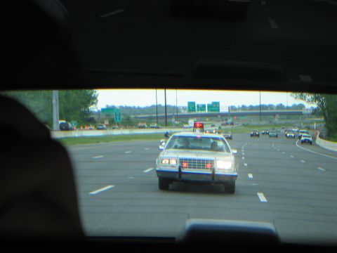 1991 Ford LTD Crown Victoria - Seen through a car's rear window, a white police car with a roof light follows on a multi-lane highway.