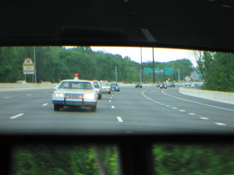 1991 Ford LTD Crown Victoria - white police car with a blue stripe and red roof light follows behind, framed by a car's rear window.