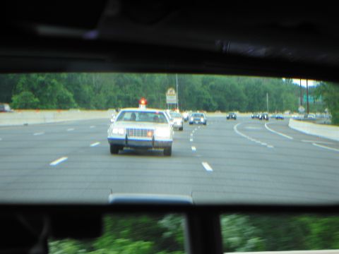 1991 Ford LTD Crown Victoria - white police car with a red roof beacon trails on a highway, viewed through the rear window of a moving car.