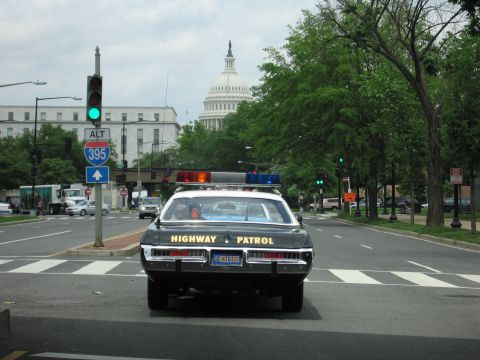 1991 Ford LTD Crown Victoria - black-and-white Highway Patrol car stops at a green light on a city street with the U.S. Capitol dome behind it.