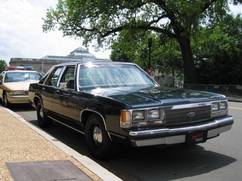 1991 Ford LTD Crown Victoria - dark Ford LTD Crown Victoria sedan with a DARE plate sits at a curb next to a tan police car under trees.