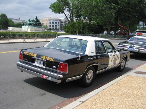 1991 Ford LTD Crown Victoria - Rear quarter of a black-and-white Ford Highway Patrol sedan parked at a curb near a memorial and other cruisers.