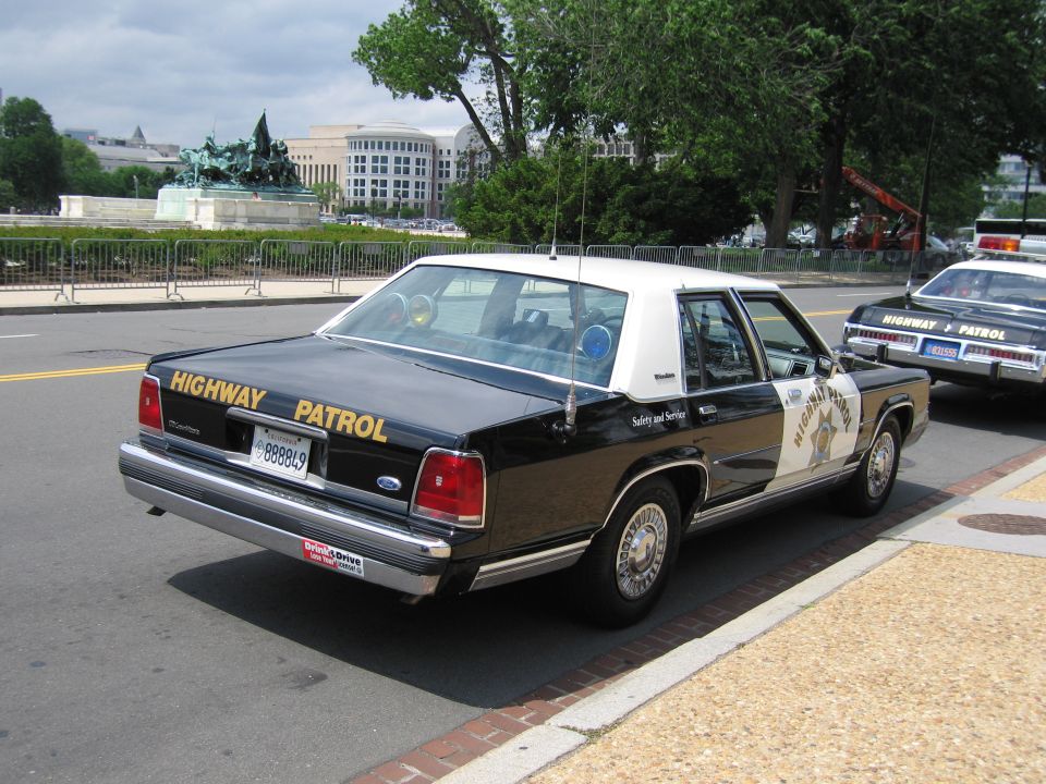 Rear of a black-and-white Highway Patrol Ford sedan parked near a monument
