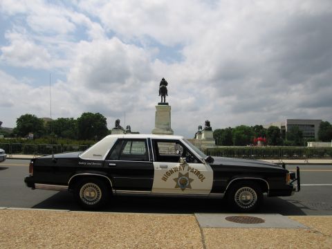 1991 Ford LTD Crown Victoria - Side view of a black-and-white Ford Highway Patrol sedan with a star emblem, parked before an equestrian statue.