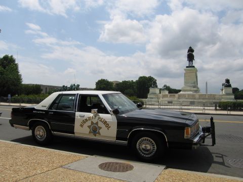 1991 Ford LTD Crown Victoria - black-and-white Ford Highway Patrol sedan with a star door emblem parked at a curb near an equestrian statue.