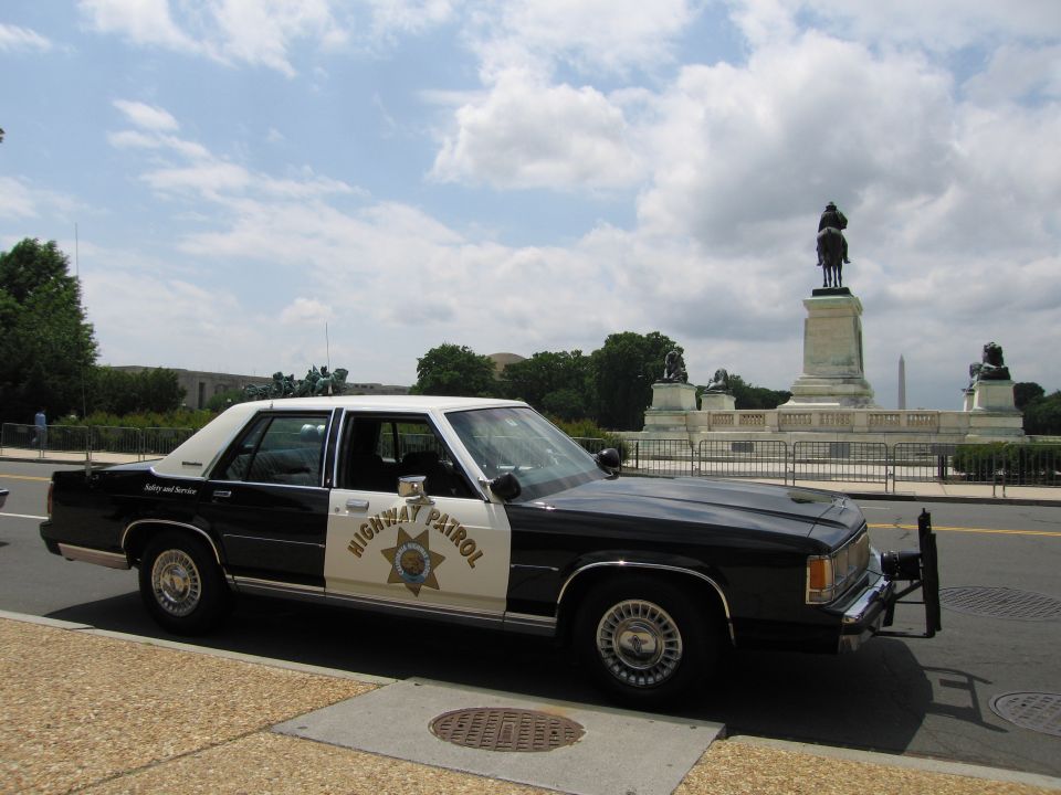 Black-and-white Highway Patrol Ford sedan parked by an equestrian monument