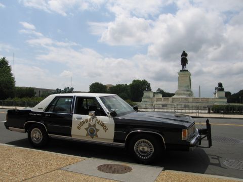 1991 Ford LTD Crown Victoria - black-and-white Highway Patrol sedan with a front push bar parked at a curb beside a memorial and statue.