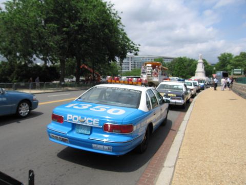 1991 Ford LTD Crown Victoria - blue Police car marked 1350 parks in a line of police cruisers along a curb near a monument and crowd.