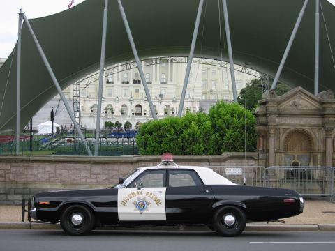 1991 Ford LTD Crown Victoria - Side profile of a black-and-white Highway Patrol four-door sedan parked before a green canopy and stone fountain.