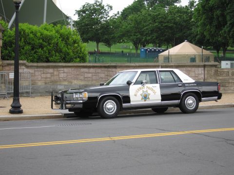 1991 Ford LTD Crown Victoria - black-and-white Highway Patrol Ford sedan fitted with a front push bar sits along a curb near a stone wall.