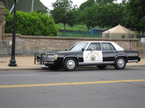 1991 Ford LTD Crown Victoria - black-and-white Highway Patrol full-size sedan with a front push bar parks curbside beside a stone wall and lawn.