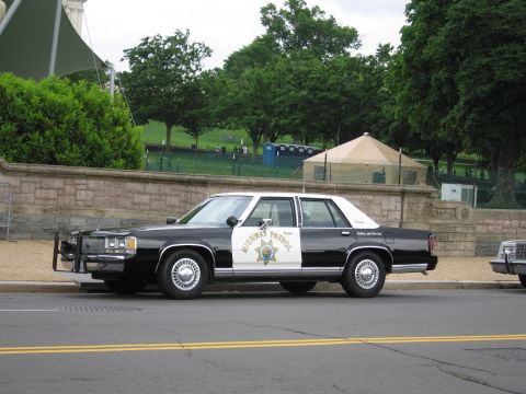 1991 Ford LTD Crown Victoria - black-and-white Highway Patrol sedan with front push bar sits curbside before a stone wall and portable toilets.