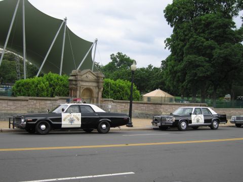 1991 Ford LTD Crown Victoria - Two black-and-white Highway Patrol full-size sedans sit nose to tail along the curb near a green canopy.