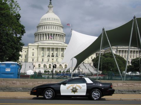 1991 Ford LTD Crown Victoria - black-and-white Highway Patrol Chevrolet Camaro sits curbside before a white canopy and the U.S. Capitol dome.