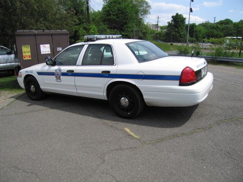 1991 Ford LTD Crown Victoria - white police sedan with a blue stripe and 'POLICE' lettering parked in a paved lot near a dumpster and trees.