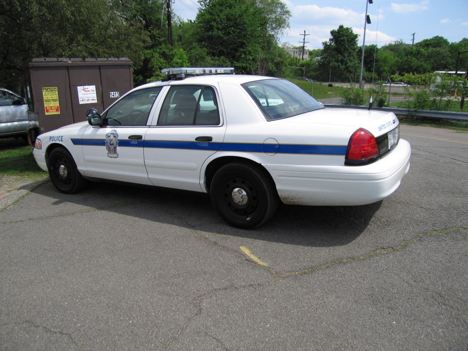 White police sedan with blue stripe parked in a lot, rear three-quarter view