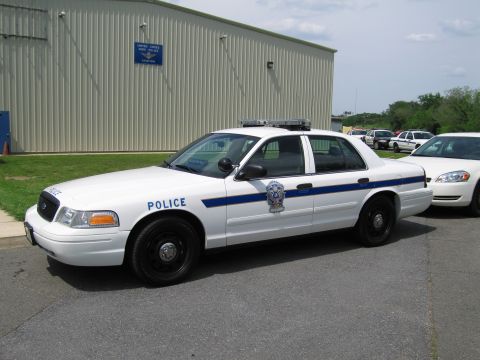 1991 Ford LTD Crown Victoria - white police cruiser with a blue stripe and shield decal parked on pavement beside a tan metal building and other vehicles.