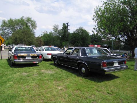 1991 Ford LTD Crown Victoria - Several police sedans, including a dark Highway Patrol car and a white cruiser, parked on grass at an outdoor gathering.