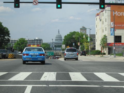1991 Ford LTD Crown Victoria - blue police cruiser stopped at a city intersection with traffic lights and a distant white domed capitol building.