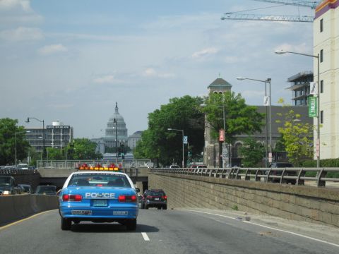 1991 Ford LTD Crown Victoria - blue police cruiser drives along a sunken roadway with a distant white domed capitol building visible ahead.