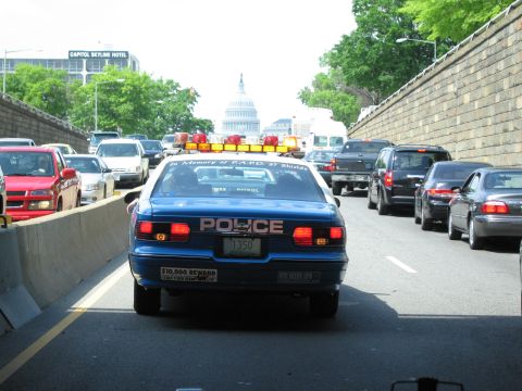 1991 Ford LTD Crown Victoria - Rear view of a blue police cruiser with memorial lettering in highway traffic, a domed capitol building visible ahead.