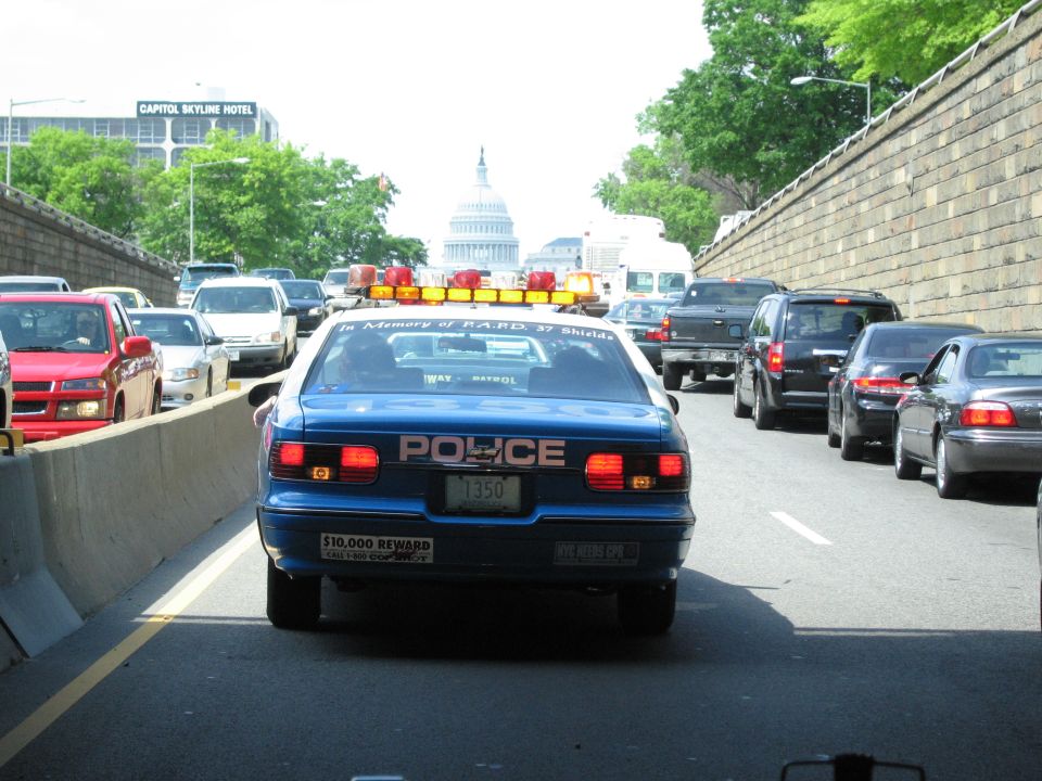 Rear of blue NYC police car in highway traffic with a distant domed capitol