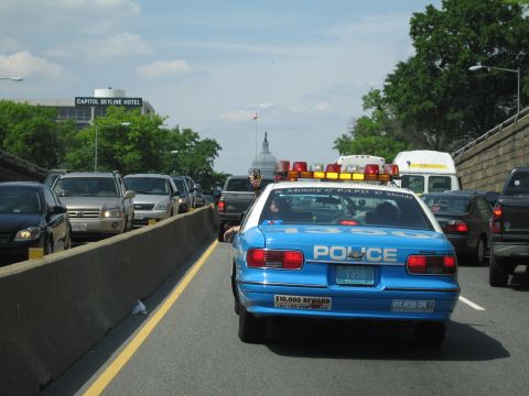 1991 Ford LTD Crown Victoria - blue police cruiser with light bar in heavy highway traffic, a distant domed capitol building and Capitol Skyline Hotel sign behind.