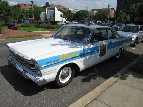 1991 Ford LTD Crown Victoria - vintage white-and-blue full-size police sedan with whitewall tires and a single beacon parked at a city curb.