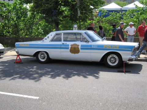 1991 Ford LTD Crown Victoria - vintage white-and-blue two-door police sedan numbered 601 with a gold badge decal on display as onlookers pass by.