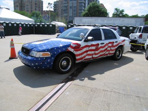 1991 Ford LTD Crown Victoria - police sedan wrapped in a stars-and-stripes American flag paint scheme parked on pavement near tents and portable toilets.