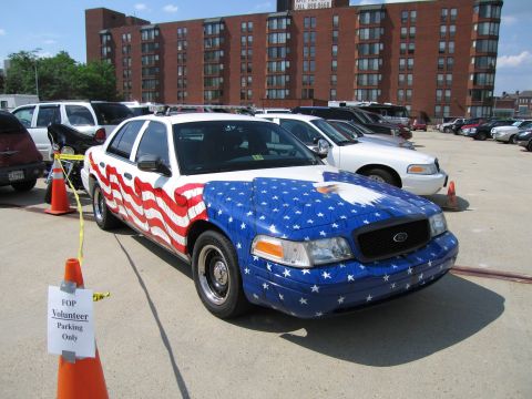1991 Ford LTD Crown Victoria - police sedan with an American flag paint wrap parked in a lot beside other cruisers in front of a tall brick building.