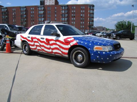1991 Ford LTD Crown Victoria - police cruiser with a flowing American flag paint scheme parked on pavement near motorcycles and a brick apartment building.