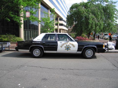 1991 Ford LTD Crown Victoria - black-and-white four-door Highway Patrol sedan with a star emblem is parked along a curb beside an office building.