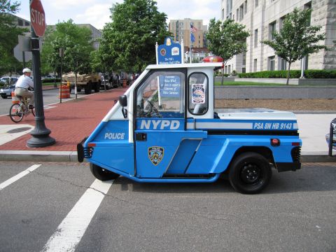 1991 Ford LTD Crown Victoria - blue-and-white NYPD three-wheeled police scooter cart with a dome light sits at a curb near a stop sign downtown.