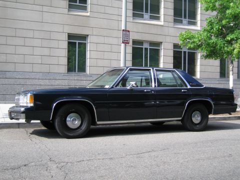 1991 Ford LTD Crown Victoria - dark blue full-size four-door sedan with steel wheels and chrome trim sits parked along a curb by a stone building.