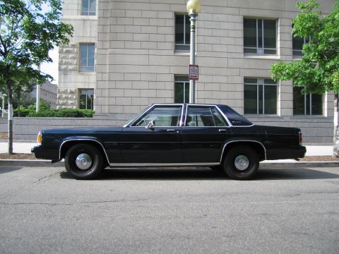 1991 Ford LTD Crown Victoria - Side profile of a dark blue full-size four-door sedan parked at the curb beside a lamppost and stone building.
