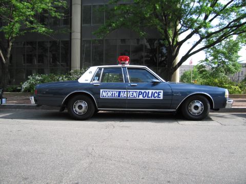 1991 Ford LTD Crown Victoria - dark gray four-door North Haven Police sedan with a red dome light is parked along a tree-lined city curb.
