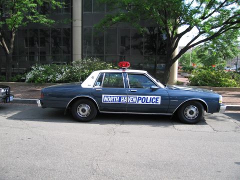 1991 Ford LTD Crown Victoria - Side profile of a dark gray North Haven Police four-door sedan with a red roof beacon parked at the curb.