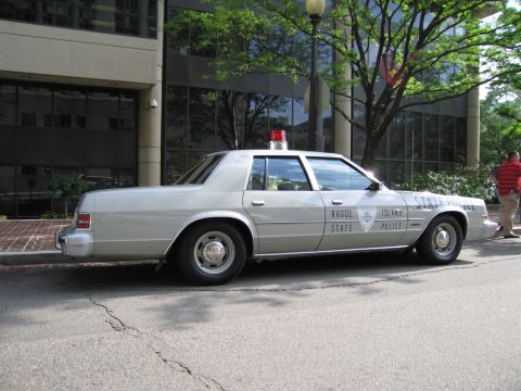 1991 Ford LTD Crown Victoria - silver Rhode Island State Police four-door sedan with a red dome light is parked along a curb by a glass building.