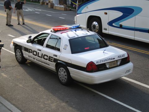 1991 Ford LTD Crown Victoria - Rear view of a black-and-white City of Alexandria Police sedan with a light bar parked along a street near a tour bus.